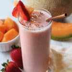 A smoothie in a tall glass garnished with strawberry & cantaloupe with cantaloupe in the background.
