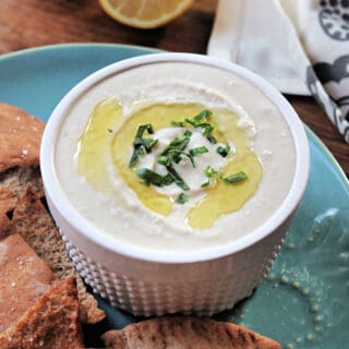 White Bean Dip in a white ramekin atop a turquoise plate with pita chips scattered around.