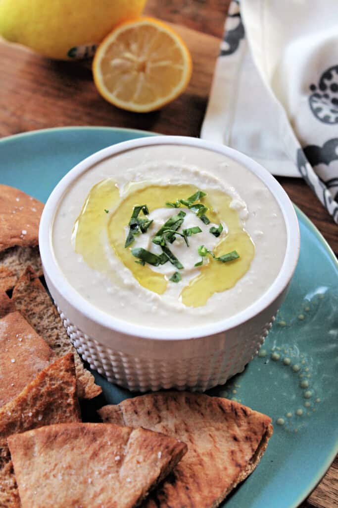 White Bean Dip in a white ramekin atop a turquoise plate with pita chips scattered around.