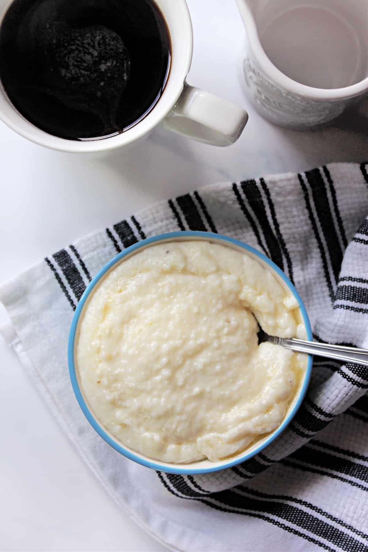 Bowl of cheese grits with a spoon on a black and white striped tea towel next to a cup of coffee..
