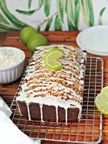 Close up of Banana-Coconut Bread with lime glaze on a cooling rack surrounded by a bowl of shredded coconut, sliced and whole limes and a kitchen towel.