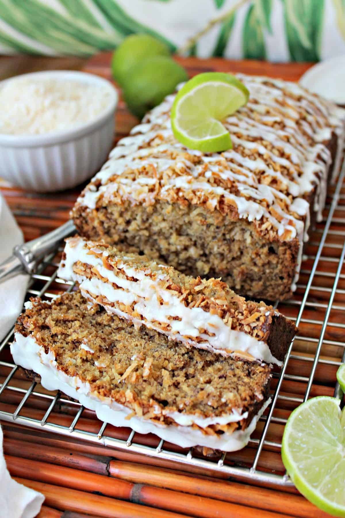 Close up of sliced Banana-Coconut Bread with lime glaze on a cooling rack surrounded by a bowl of shredded coconut, sliced and whole limes and a kitchen towel.