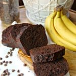 A sliced loaf of Double Chocolate Banana Bread on a wood cutting board surrounded by chocolate chips with bananas in the background.