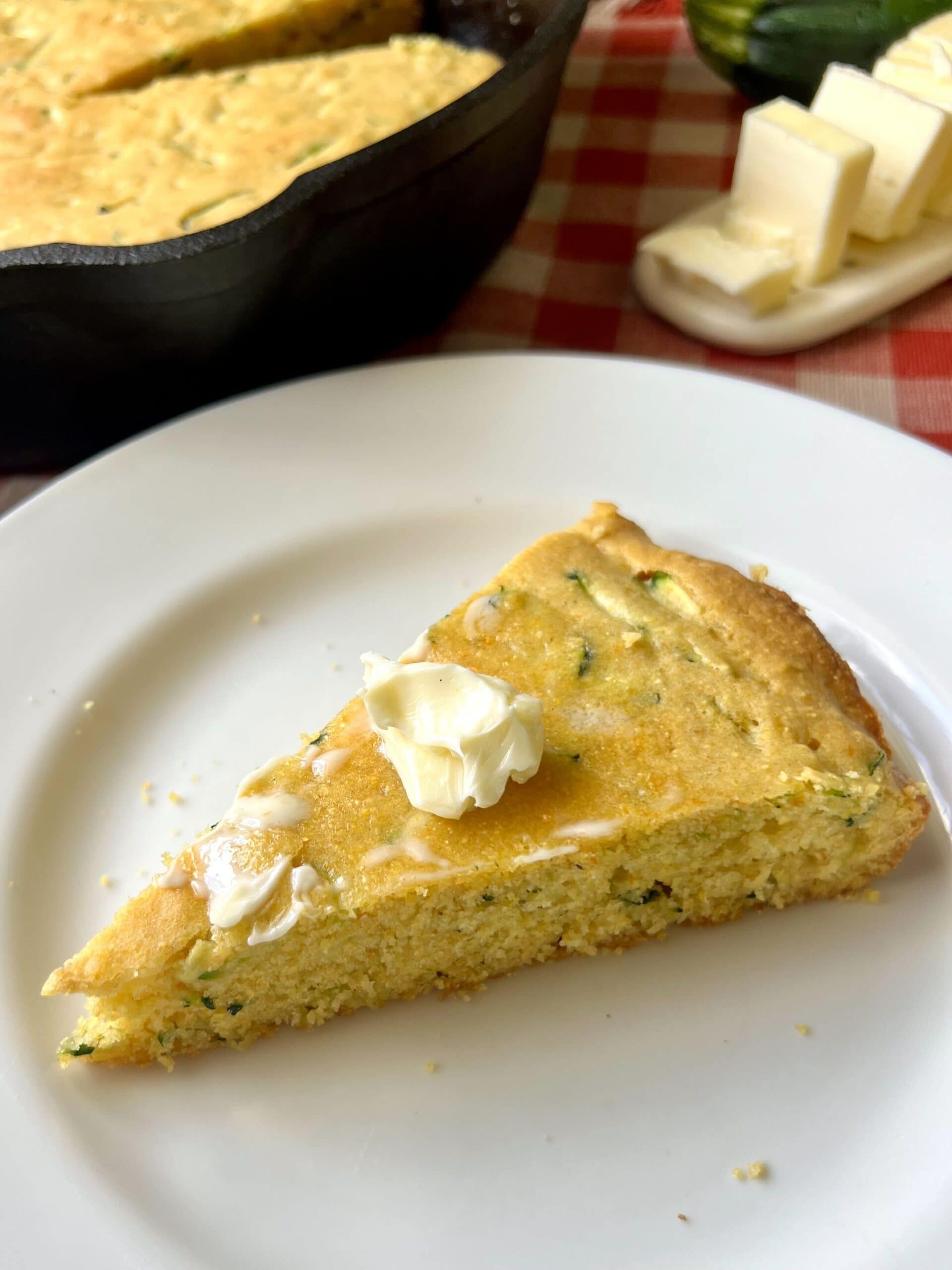 A wedge of zucchini cornbread topped with a pat of butter on a white plate.