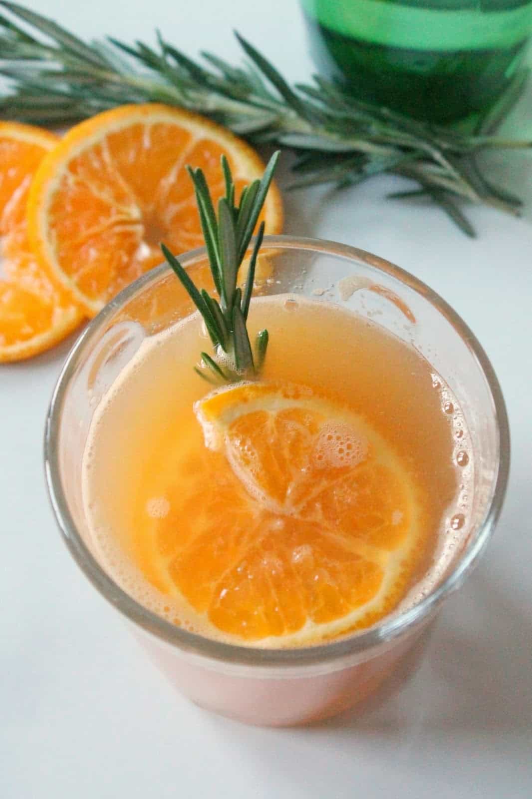 An orange mocktail in a glass with a slice of orange and sprig of rosemary.