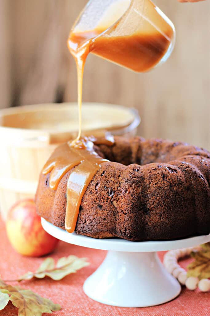 Honey Apple Bundt Cake on a white cake stand being drizzled with Honey Caramel Sauce.