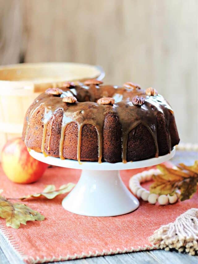 Honey Apple Bundt Cake on a white cake plate with fall leaves and apple in background.
