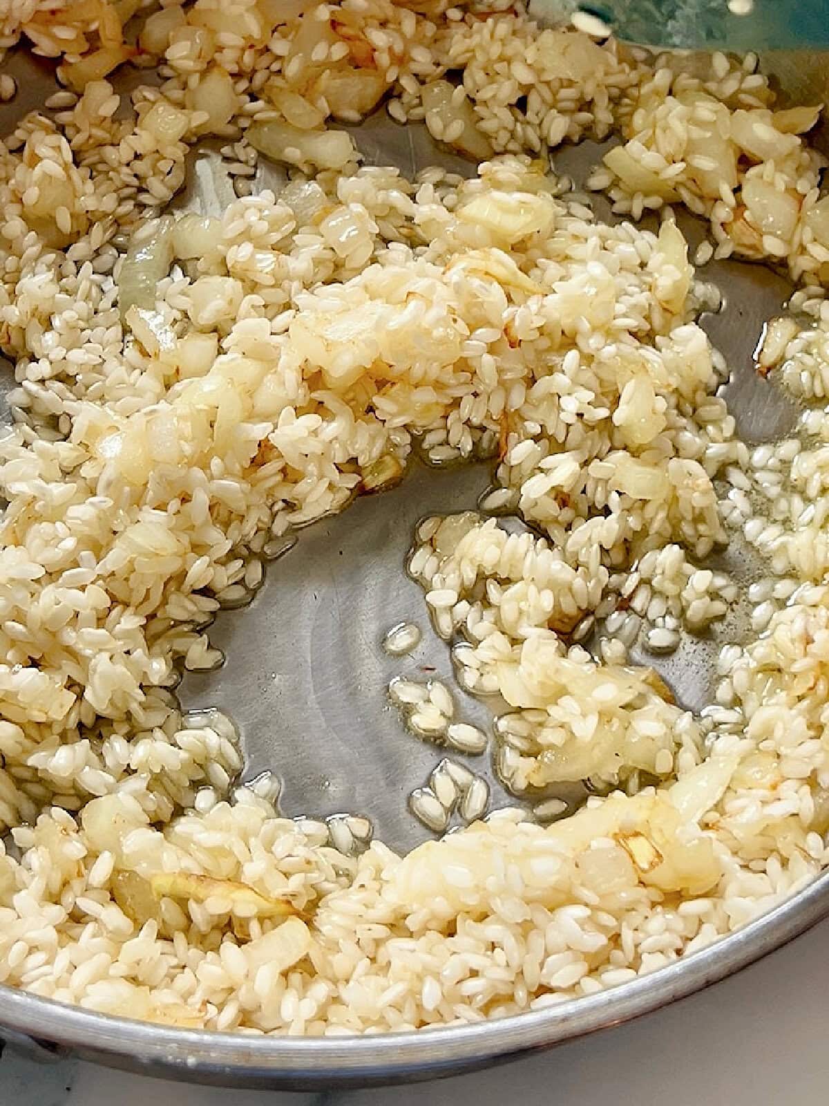 Aromatics and arborio rice being sauteed in olive oil in a pan.