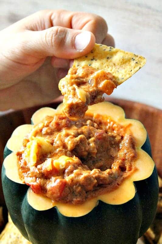 Close up of hand dipping tortilla chip into pumpkin chili cheese dip.