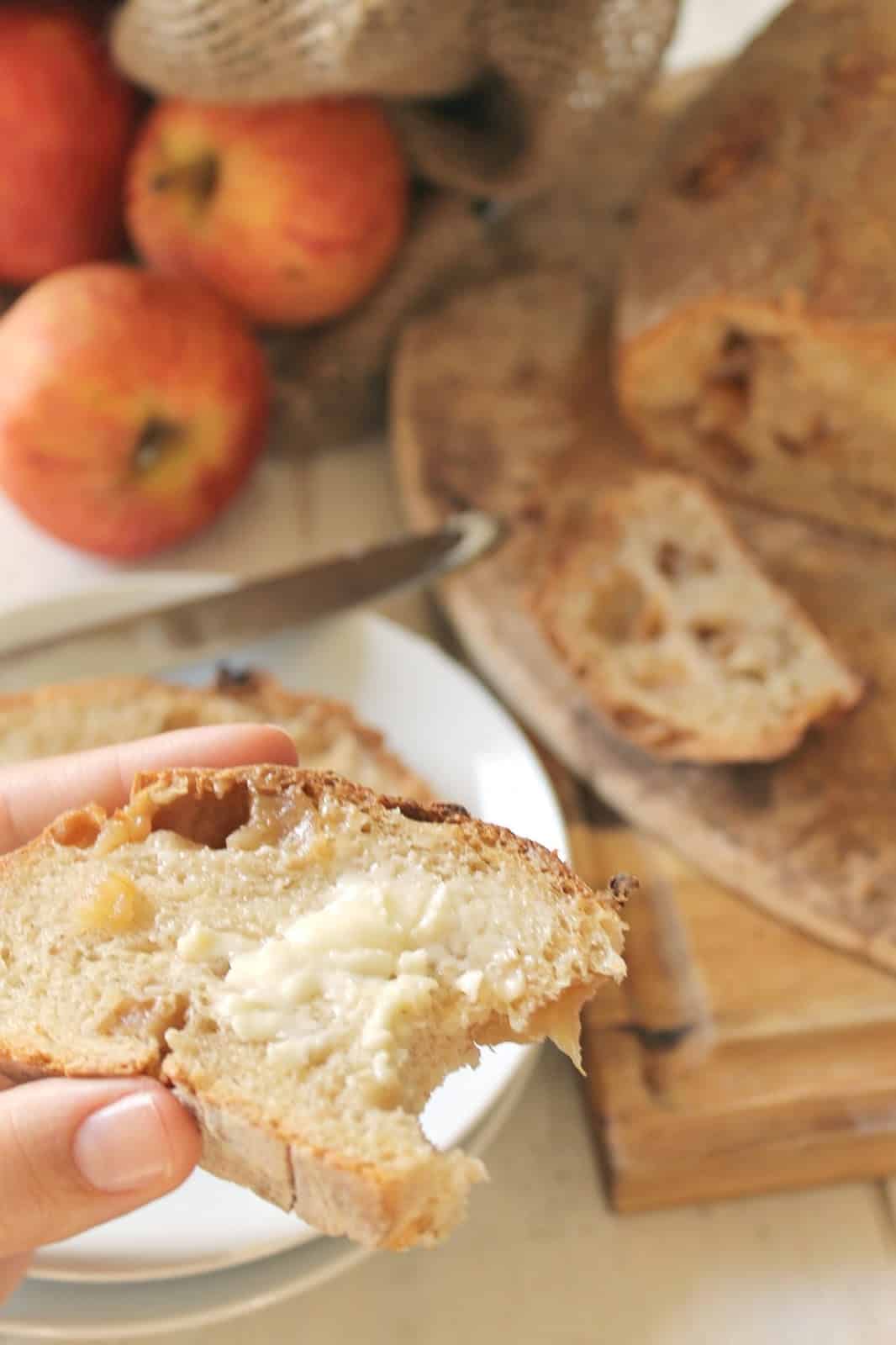 A hand holding a slice of buttered Roasted Apple Bread with a bite taken out of it.