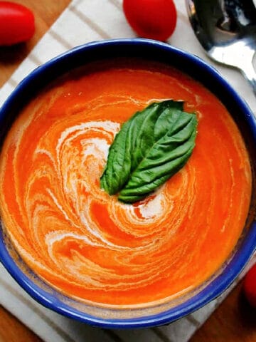 A blue bowl full of spicy tomato soup garnished with a piece of basil and a swirl of cream.