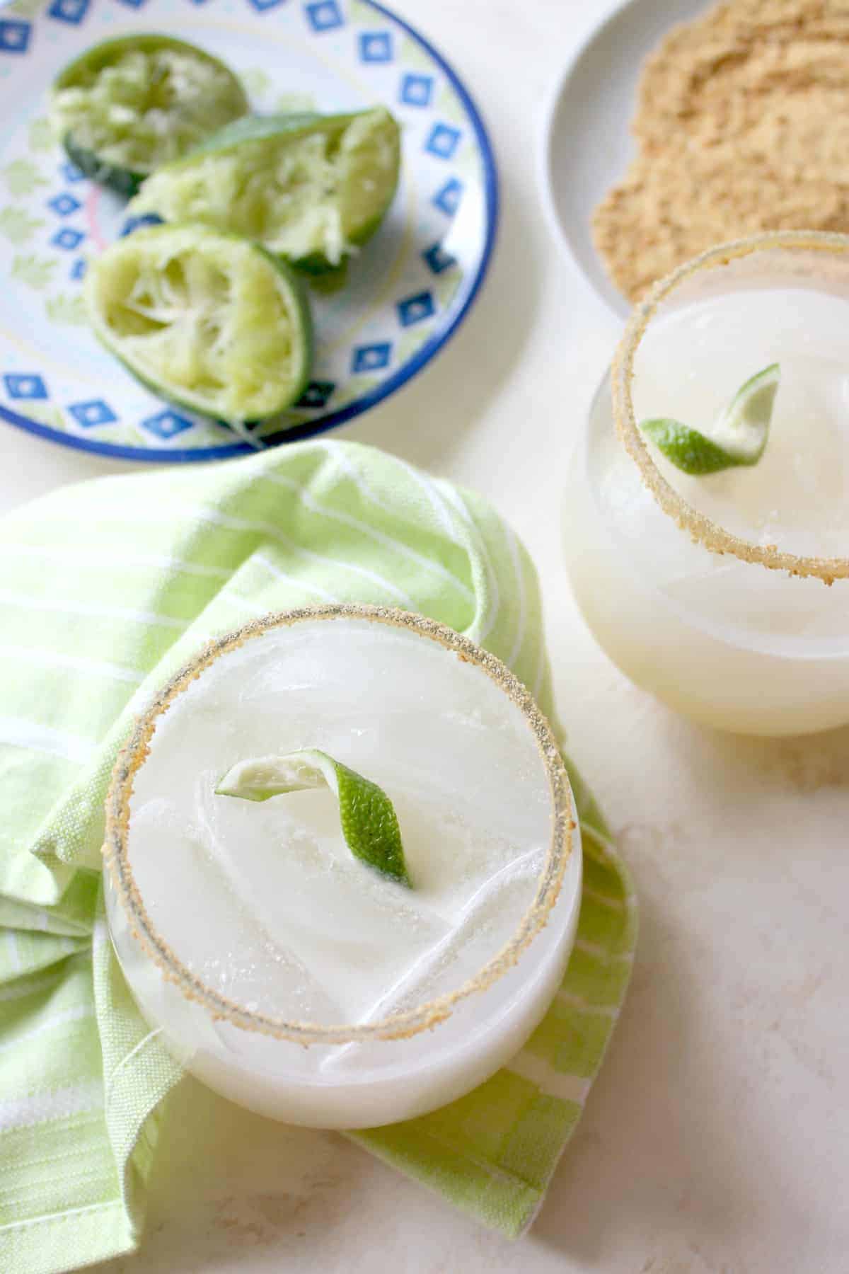 Coconut lime drinks in glasses with squeezed limes & green napkin beside them.