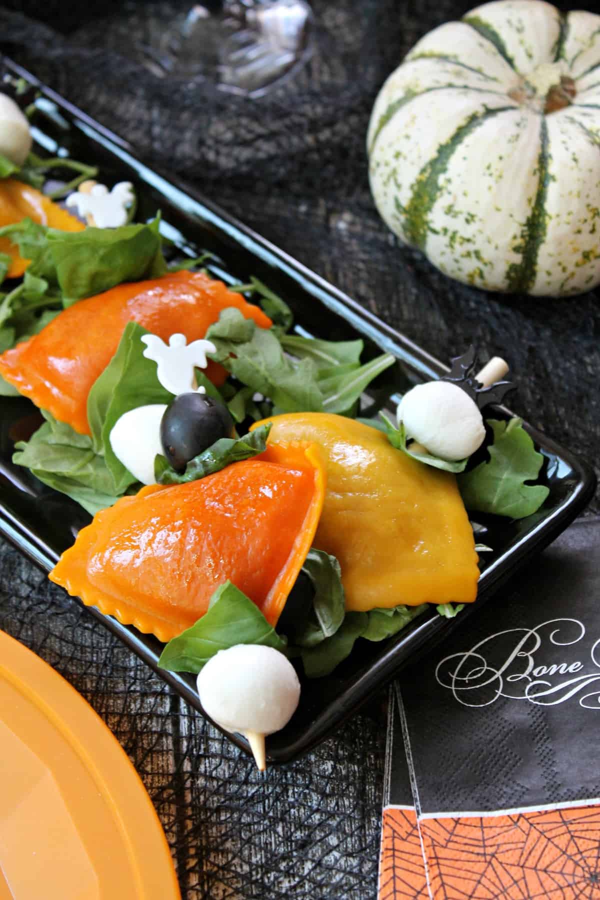 Orange and yellow pumpkin ravioli threaded onto bamboo skewers with mozzarella balls, black olives and fresh basil on a Halloween background.