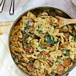 A large skillet filled with Creamy Mushroom & Chicken Sausage Florentine Pasta with a yellow and white striped dish towel underneath.