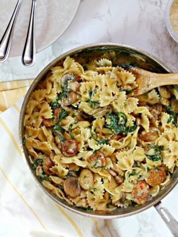 A large skillet filled with Creamy Mushroom & Chicken Sausage Florentine Pasta with a yellow and white striped dish towel underneath.