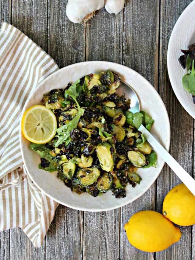 Overhead shot of Lemon Parmesan Roasted Brussels Sprouts on a porcelain plate surrounded by lemons and a striped kitchen towel.