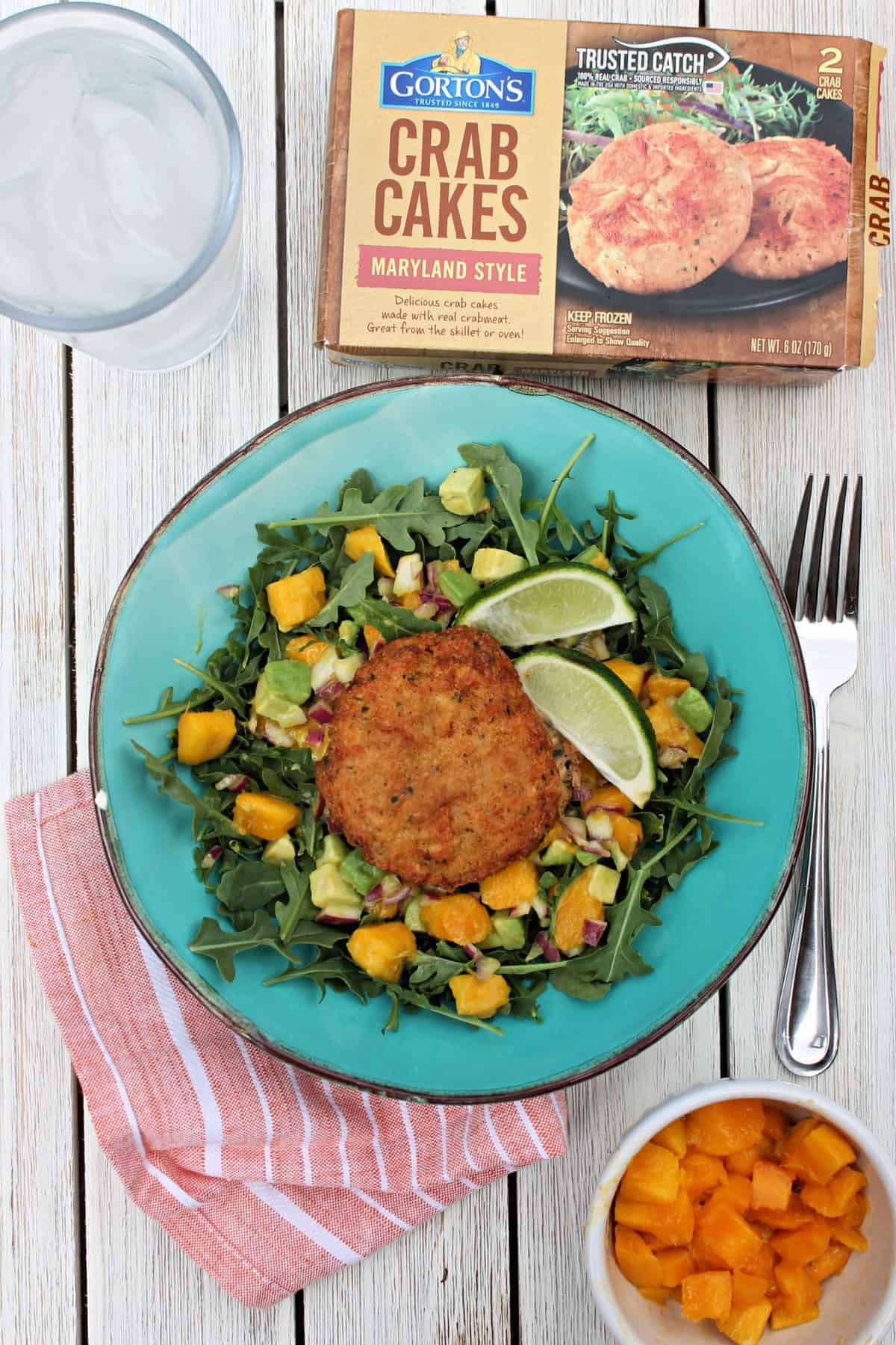 Overhead shot of Crab Cake Salad in a turquoise dish on a picnic table with a napkin, fork, water glass and product box.