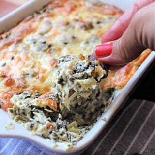 Hand holding a tortilla chip being dipped into Hot Spinach & Artichoke Dip in a white baking dish.