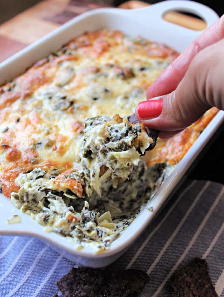 Hand holding a tortilla chip being dipped into Hot Spinach & Artichoke Dip in a white baking dish.