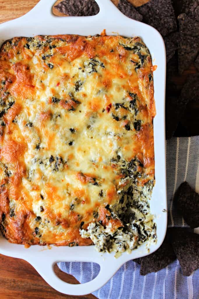 Bubbly cheese topping on Hot Spinach and Artichoke Dip in a white baking dish with blue corn tortilla chips surround it.