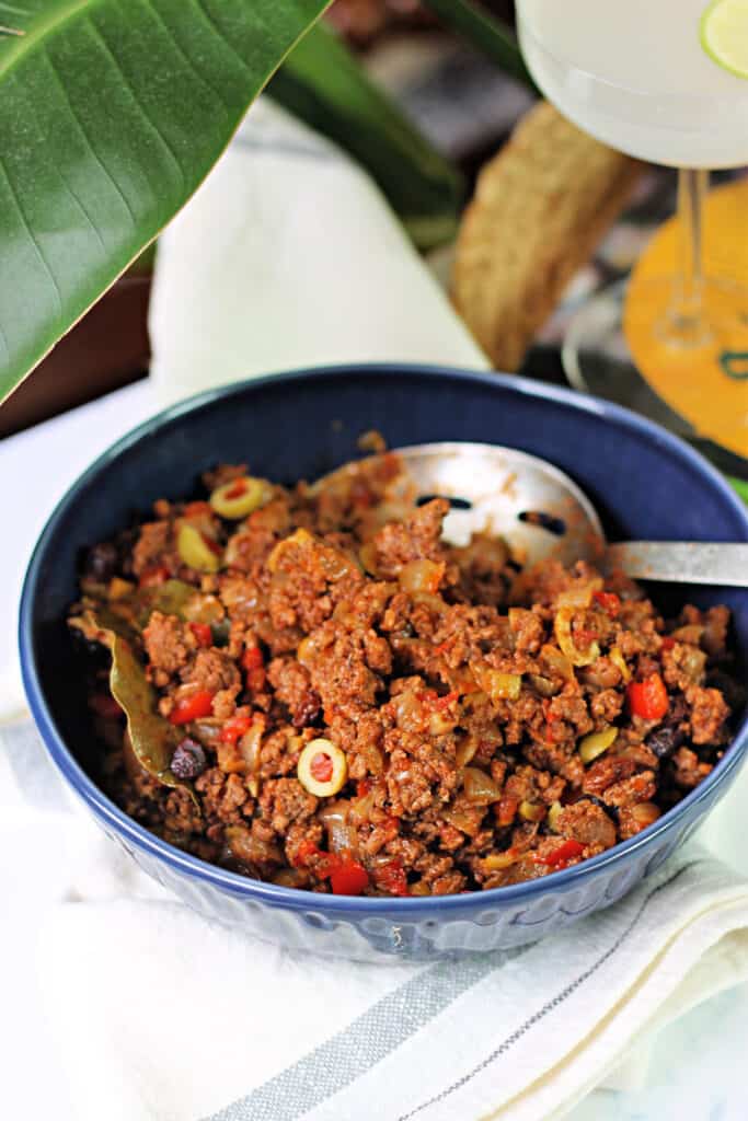 Cuban Picadillo in a blue ceramic bowl with a tropical leaf in background.