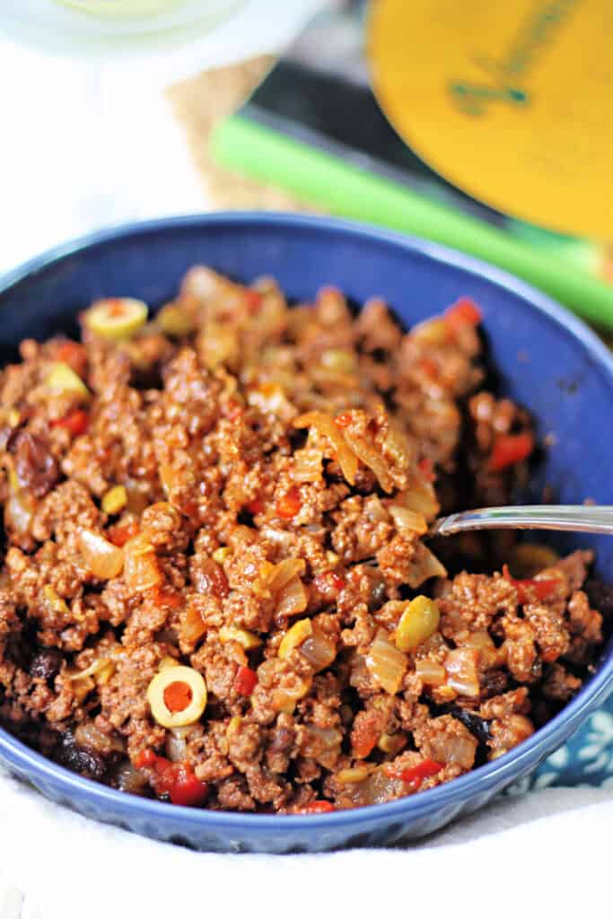 Spoon scooping Cuban Picadillo out of a blue ceramic bowl.