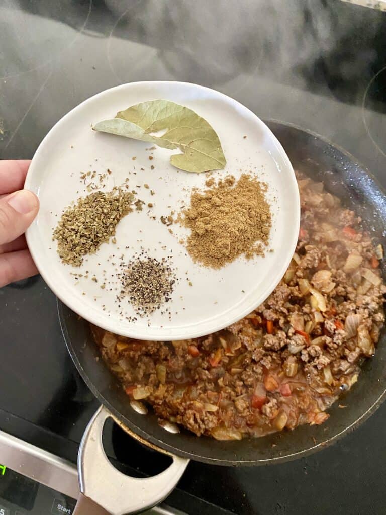 Adding bay leaf, cumin, black pepper and dried oregano to a skillet with ingredients for Cuban Picadillo.