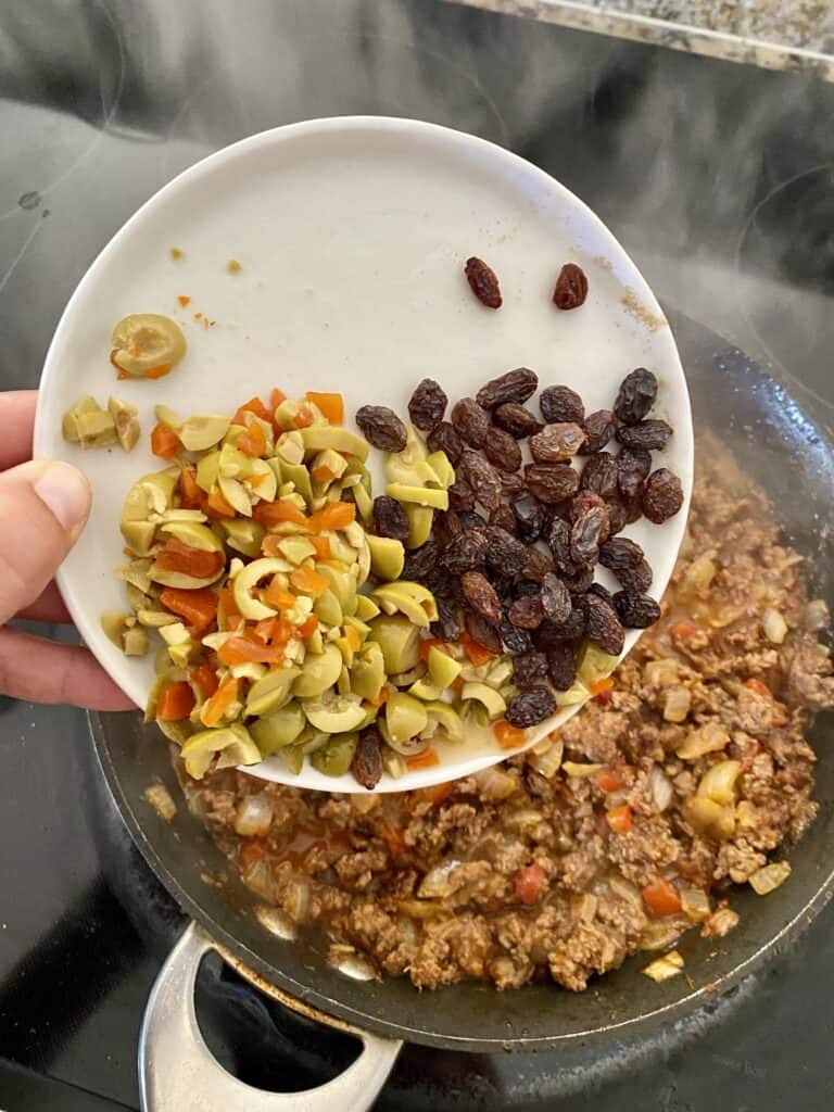 Adding chopped pimento stuffed green olives and raisins to Cuban picadillo in a skillet.