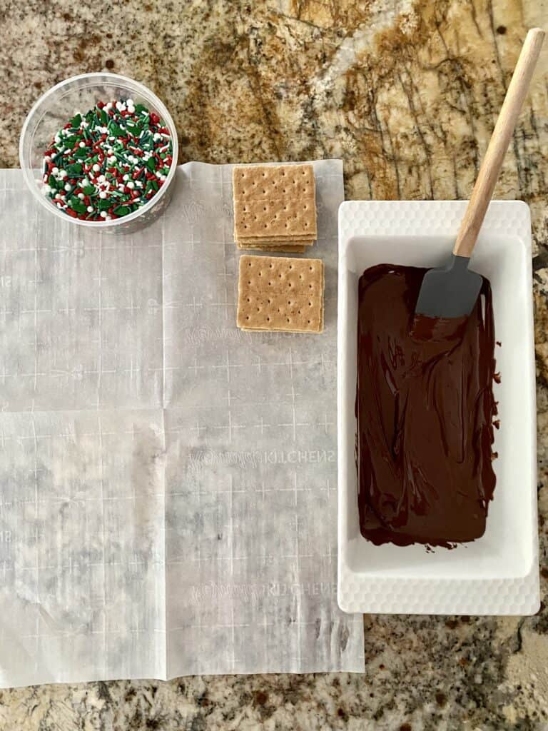 A countertop prepped with parchment paper, melted chocolate, graham crackers and sprinkles in preparation for Chocolate Covered Graham Crackers.