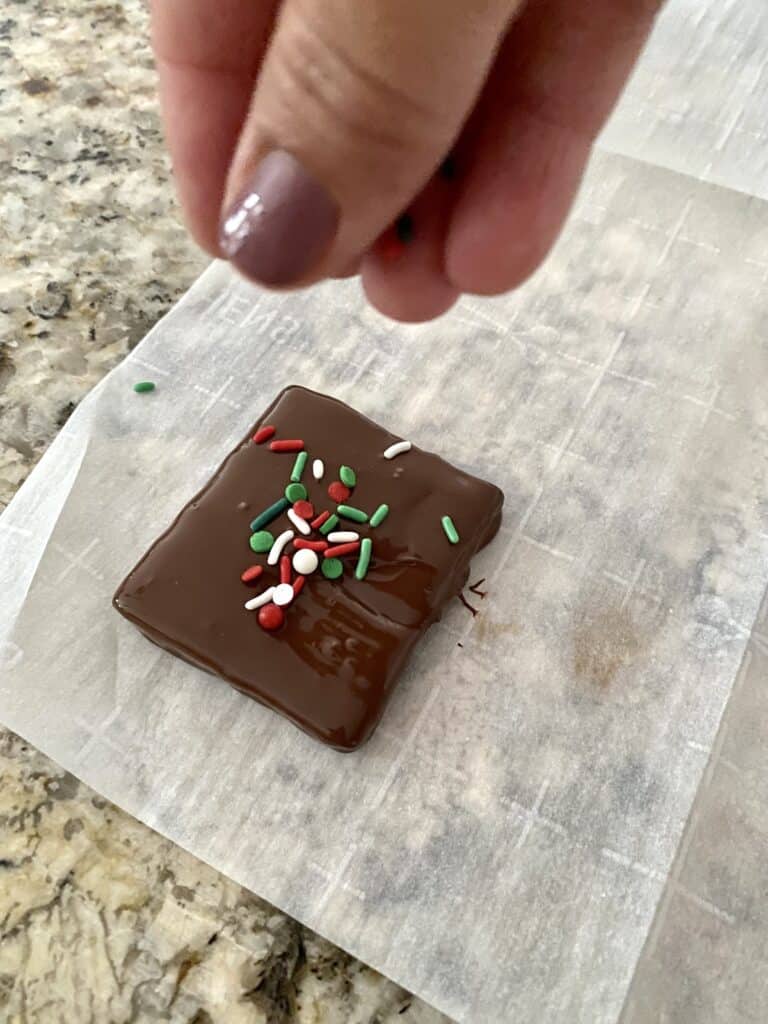 A chocolate covered graham cracker placed on a sheet of parchment paper being sprinkled with Christmas sprinkles.