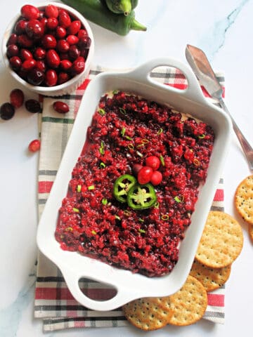 Cranberry Jalapeno Dip in a white serving dish placed on top of a green and red plaid napkin surrounded by cranberries, jalapenos & crackers.
