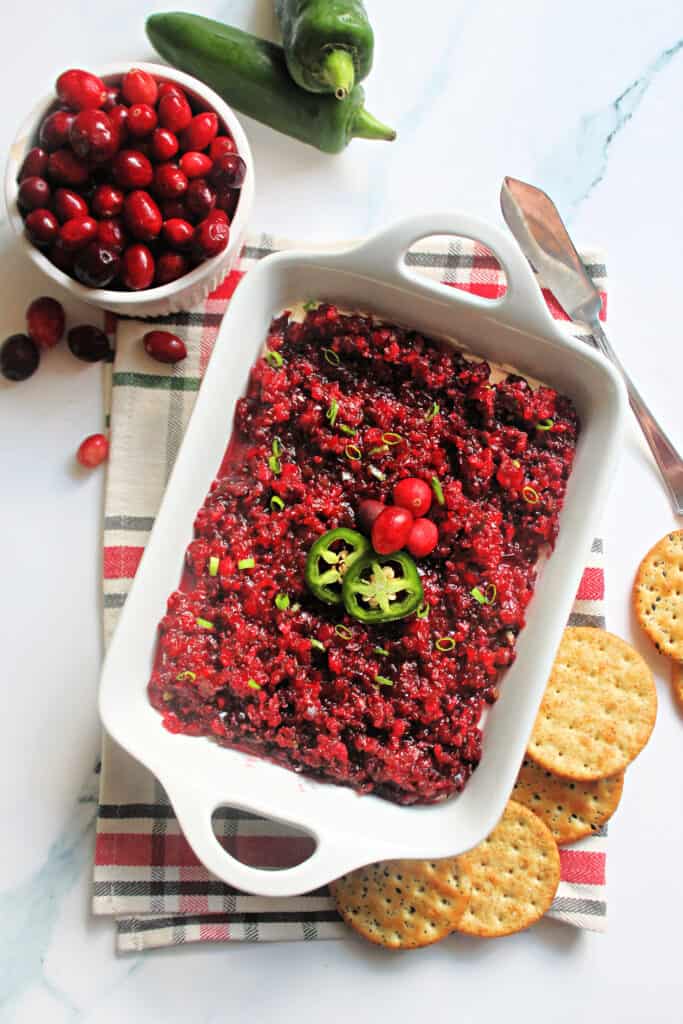 Cranberry Jalapeno Dip in a white serving dish placed on top of a green and red plaid napkin surrounded by cranberries, jalapenos & crackers.