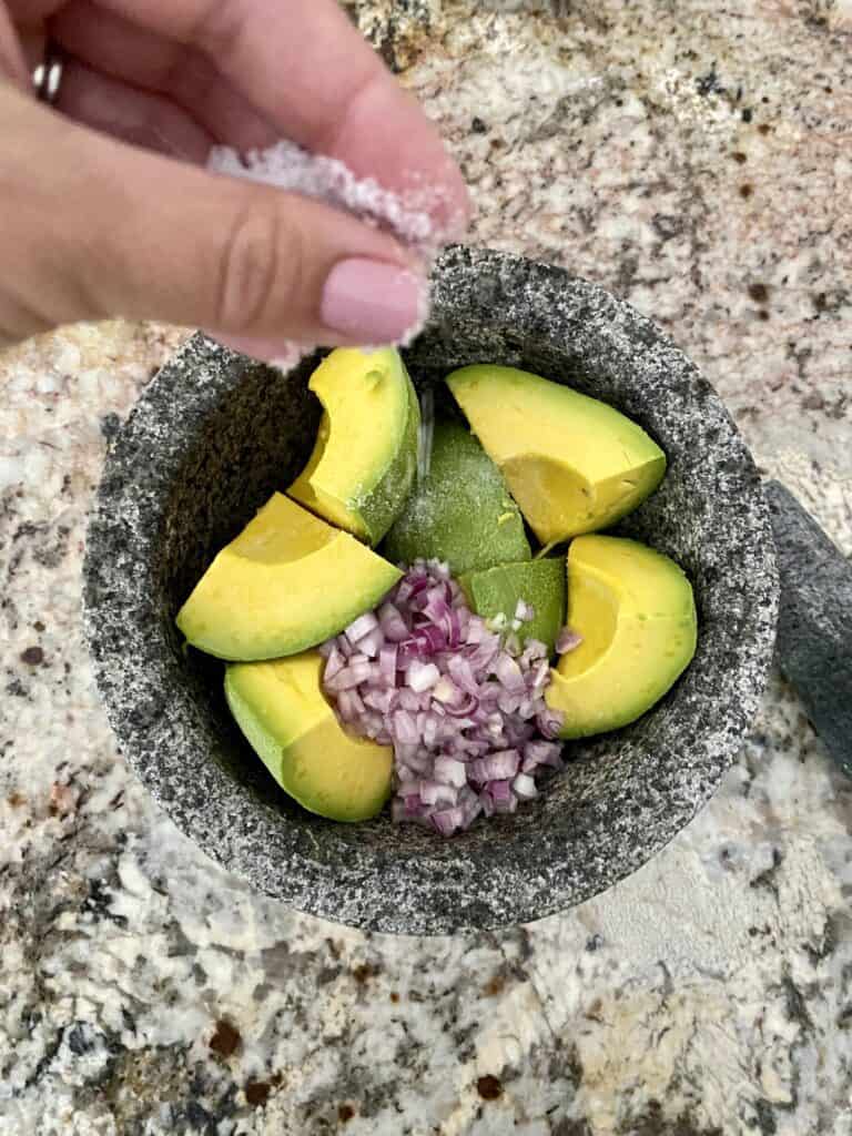 Sprinkling salt onto avocado, shallots and lime juice in a molcajete.
