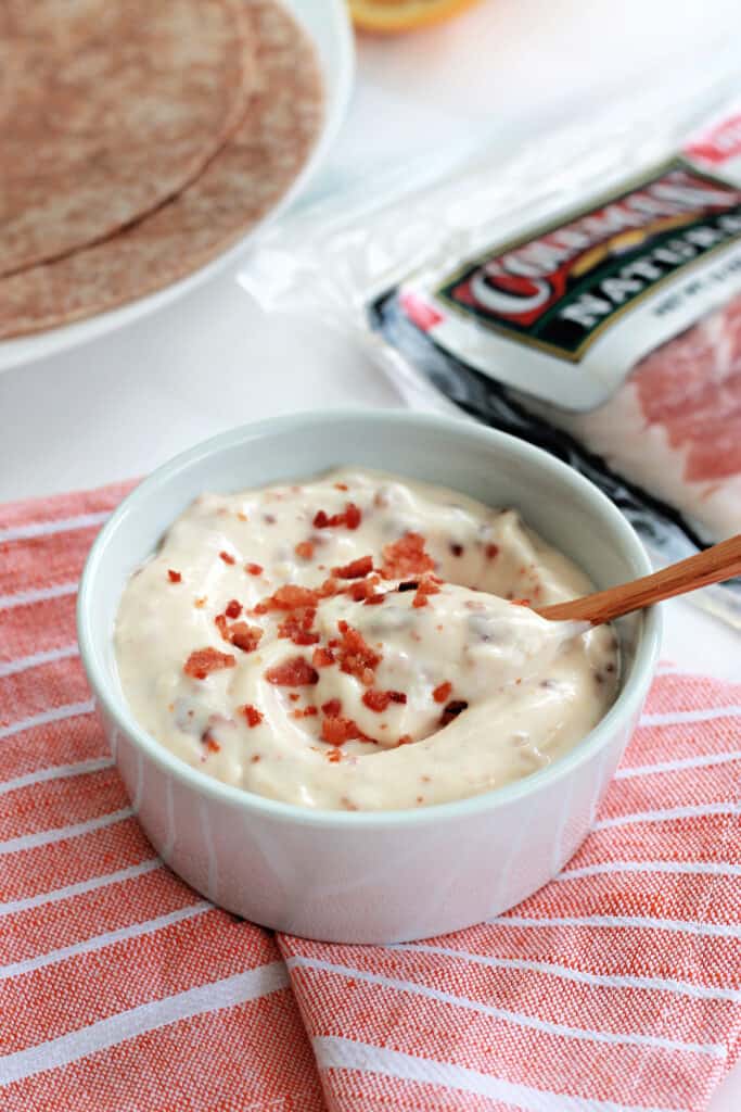 A closeup of bacon aioli in a white ramekin with a small wooden spoon scooping some out.