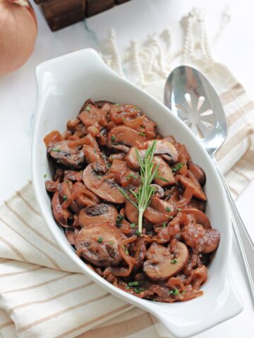 Caramelized onions and mushrooms in a white dish with a silver serving spoon beside it.