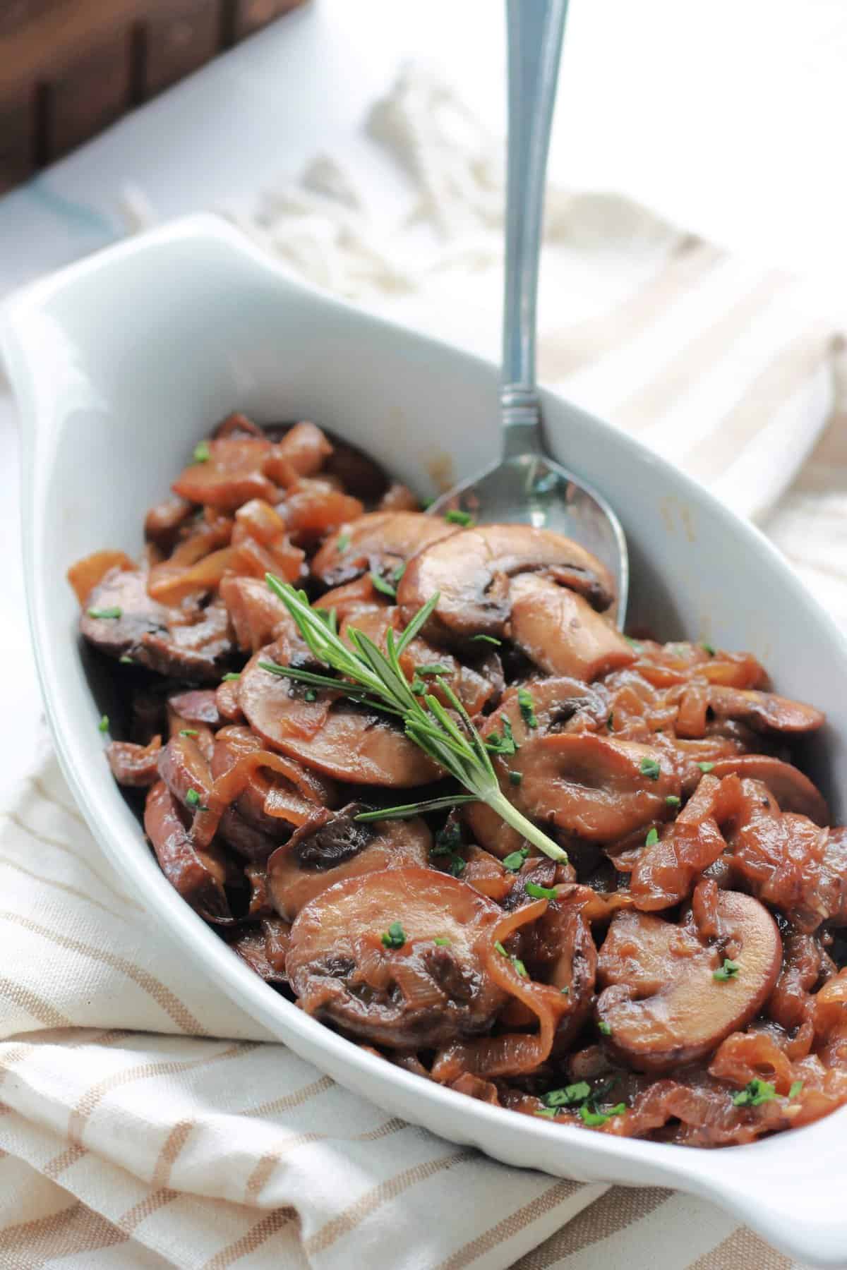 A closeup of Caramelized Mushrooms and Onions in a white dish with a sprig of rosemary on top.
