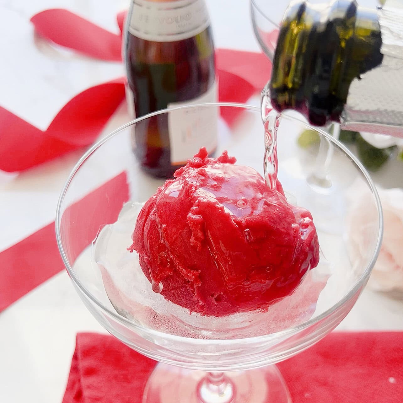 Pouring prosecco over a scoop of raspberry prosecco in a champagne coupe.