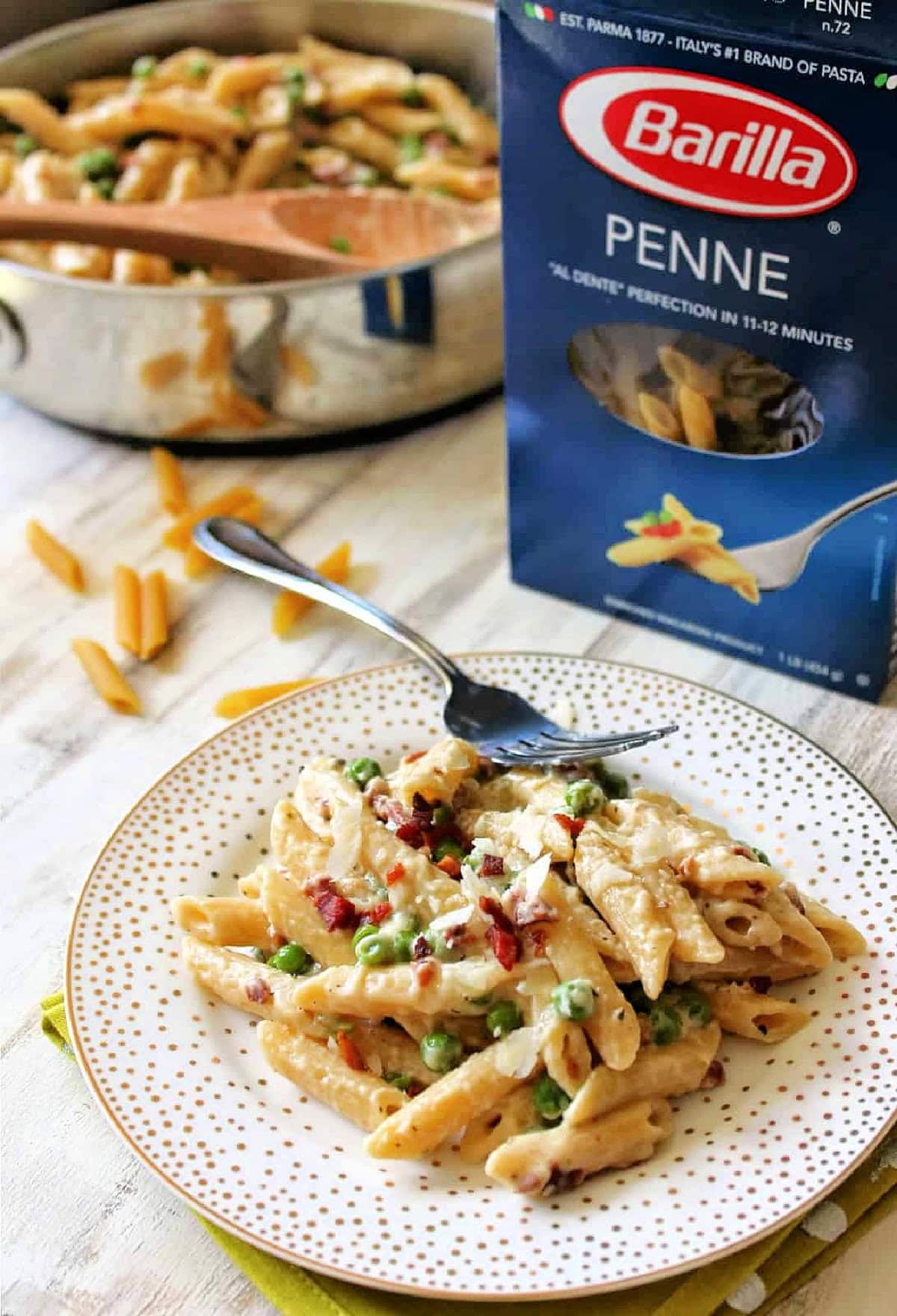 A serving of Creamy Penne Pasta with Pancetta and Peas on a plate with a box of Barilla Penne pasta in the background.